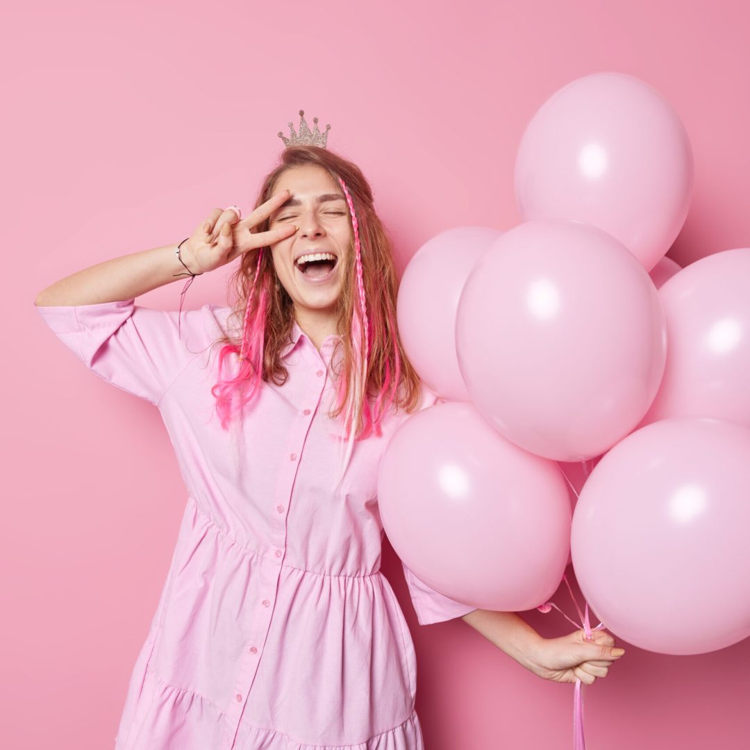Happy overjoyed birthday girl spends free time on party holds bunch of inflated balloons makes peace gesture wears crown and dress isolated over pink background laughs gladfully. Celebration concept