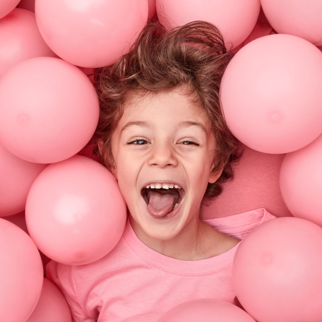 From above happy little boy showing tongue and looking at camera while playing in heap of pink balloons