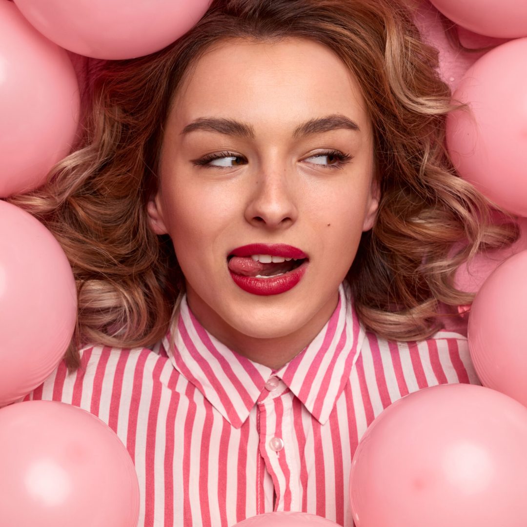 From above attractive young female licking lips and looking away while lying amidst pink balloons during party