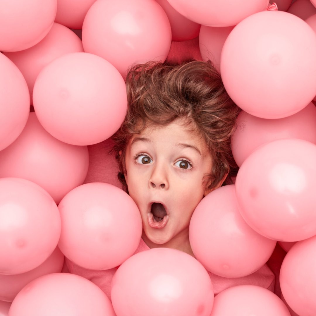 From above astonished little boy looking away while having fun under heap of pink balloons during party