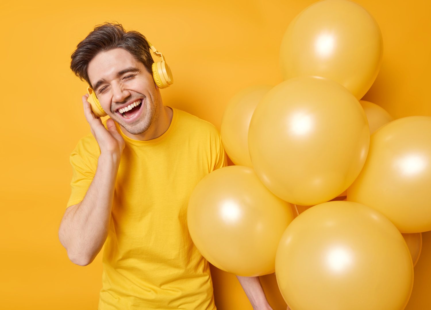 People holidays celebration and fun concept. Happy brunet adult man enjoys favorite melody keeps hand on headphones poses with helium balloons dressed casually isolated over yellow background.